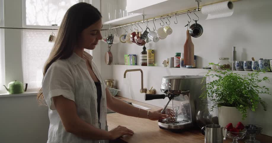 A woman in a white blouse stands at a kitchen counter and makes an espresso