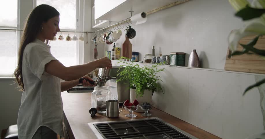 A woman wearing a white blouse stands at a kitchen counter preparing coffee