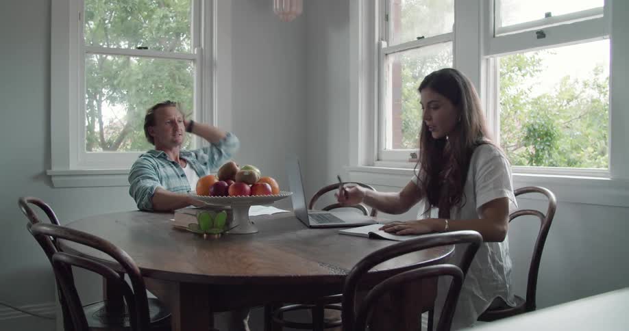 A man and woman sit at a table in a home and look at papers and a computer and talk