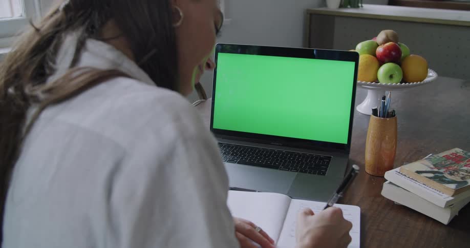 Young woman sitting at the dinner table in the kitchen at home takes notes from a laptop