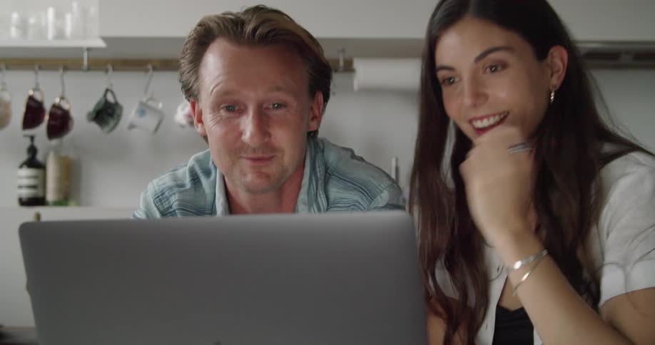 A couple laugh and wave while sitting in their kitchen and using a laptop for a video call