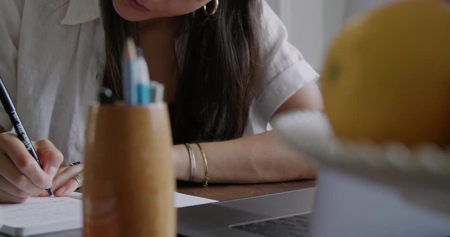 A woman glances at the screen of a laptop before writing in a notebook while sitting at a table indoors during the day