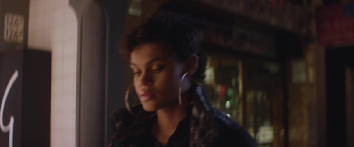 A woman with short hair and big hoop earrings walking in the city in front of shops