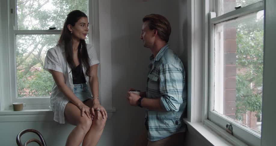 A man leaning against a wall chats with a woman sitting on the sill of a window in a house during the day