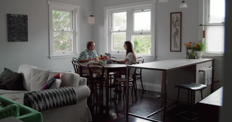 A man chats with a woman as they two sit together at a dining table in a house during the day