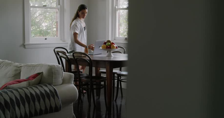A woman sits at a table drinking a beverage while working on her laptop
