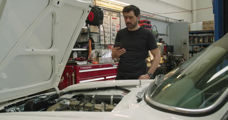 Man scrolls through a smartphone while standing in front of a white vehicle with its hood up inside of an automotive workshop