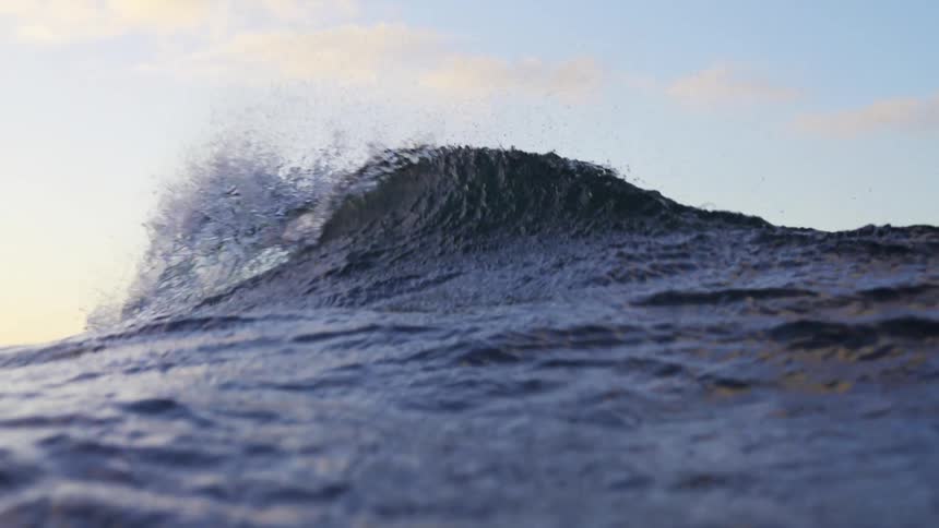 A wave crests in the ocean with a blue sky and clouds visible