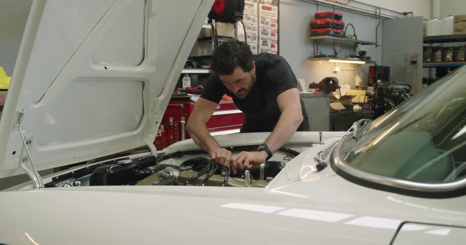 A male mechanic has a car hood open and is assessing hoses on the engine of a car in a garage
