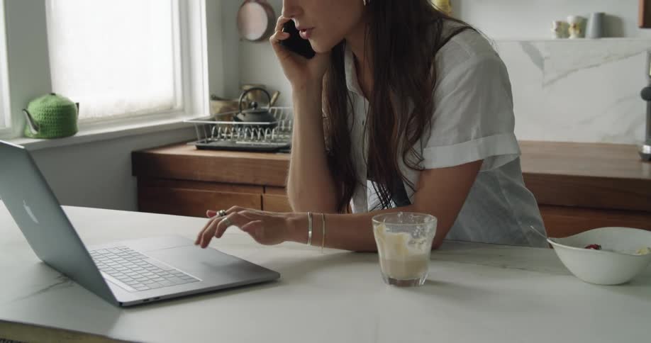 A woman speaks on a smartphone while typing on a laptop in a home kitchen during the day