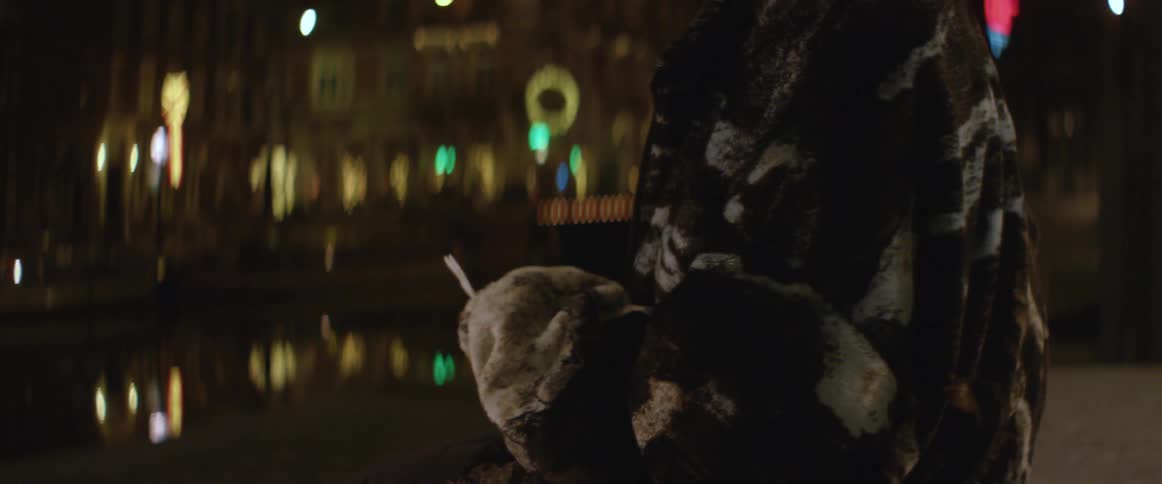 A woman standing by the water in a city at night looks around and then opens up a note and reads it