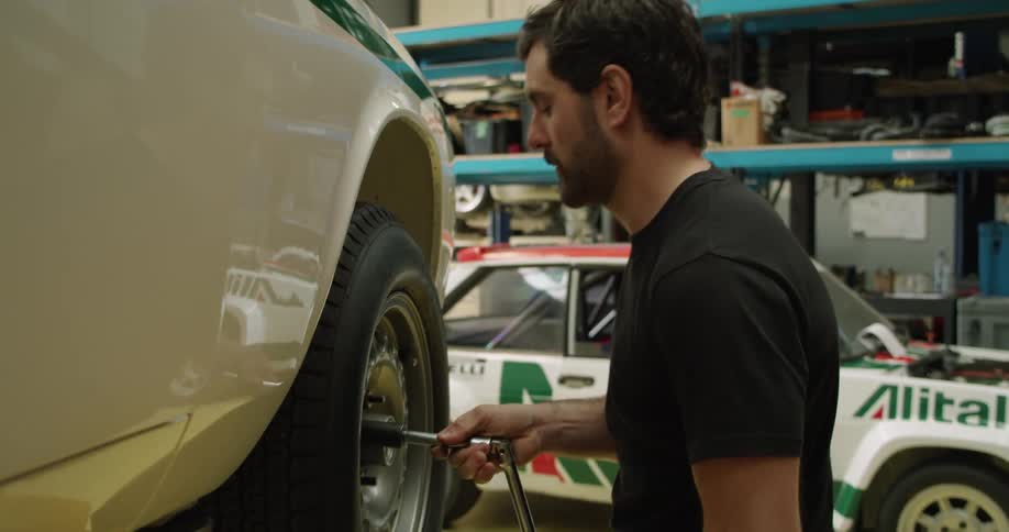 Man with a tire iron inside of a mechanic shop taking a tire off of a white car lifted into the air