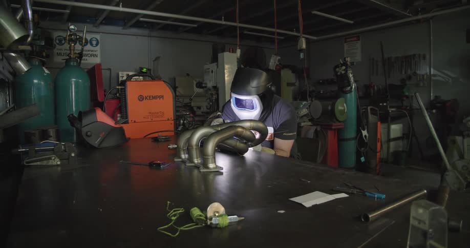 A worker with a solder mask soldering engine exhaust pipes in a dimly lit workshop