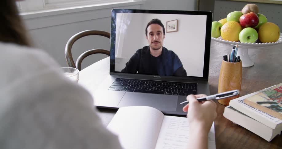 A woman sits at a dining table writing as she talks with a man on her laptop