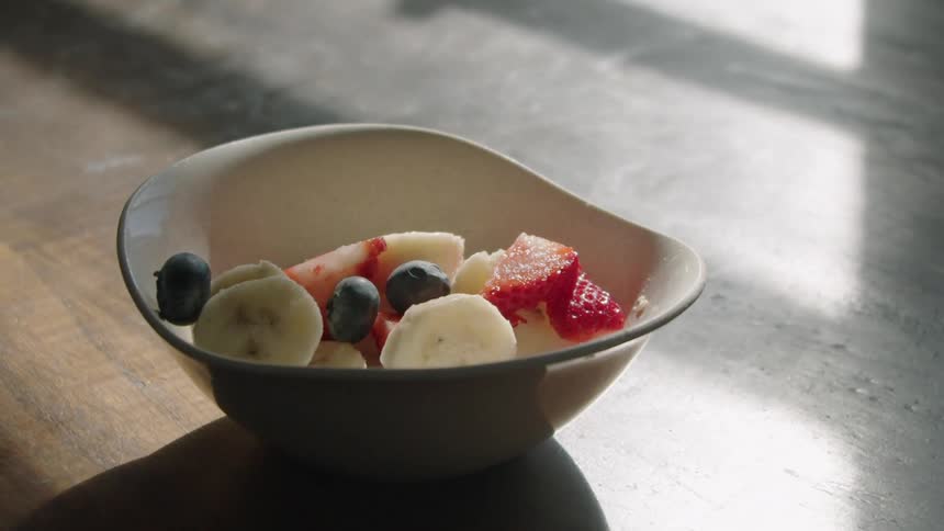 Blueberries dropping into a bowl of cut bananas and strawberries on a wooden surface