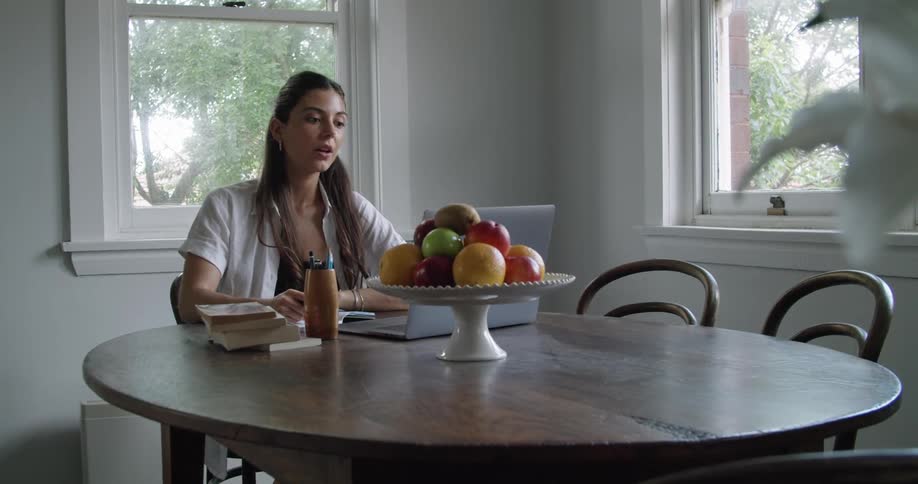 A woman drinks from a glass of water before writing in a notebook as she participates in a video chat using her laptop