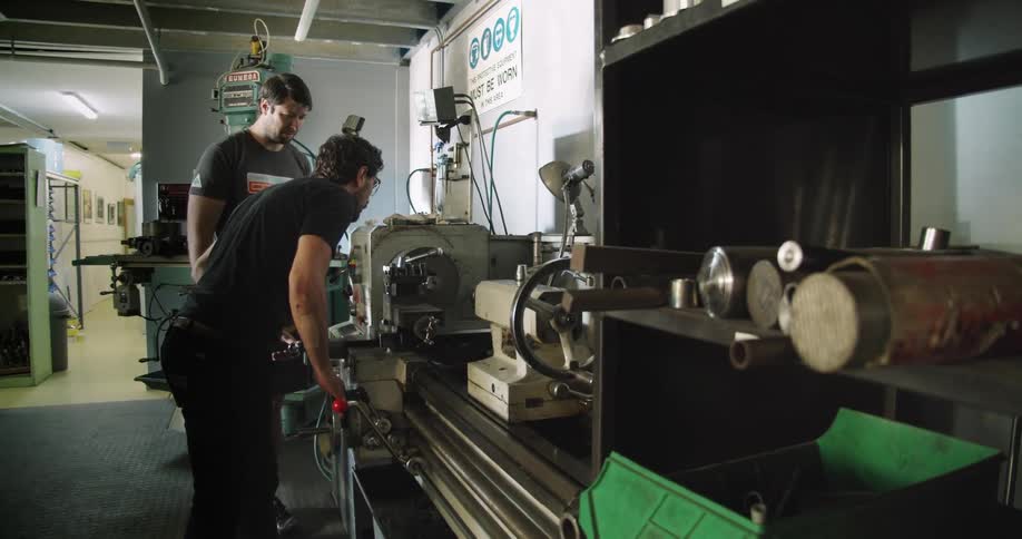A pair of men without safety equipment lean forward to closely watch a lathe as they operate it below a sign stating that safety equipment is required
