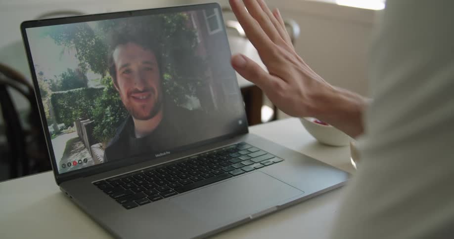 A caucasian man video chats on his computer inside his home with another caucasian man who is outside they smile wave and give each other a thumbs up