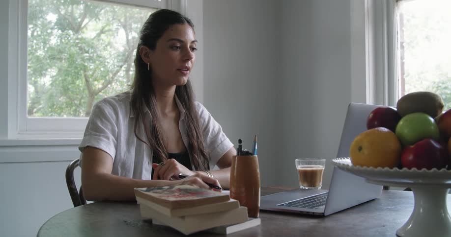 Woman receiving online classes at home through a laptop on the dinning table