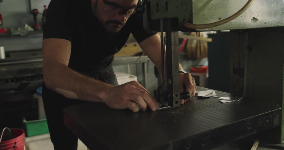A man in a workshop examines a piece of metal and then places it under a large tool