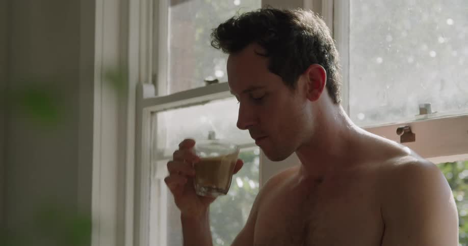 A bare chested man drinks coffee and looks at his phone while standing inside