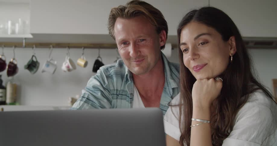 A man places his arm around his wifes shoulders as they wave and a kiss at a person during a video call on a laptop