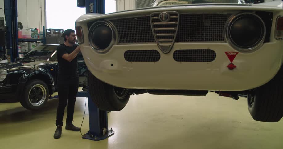 A male mechanic lowers a white car to the cement floor on a lift in a garage