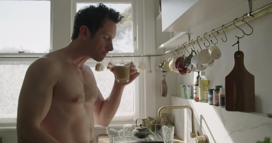 A shirtless young man sips from a glass while standing in front of a kitchen faucet before turning and carrying the drink away