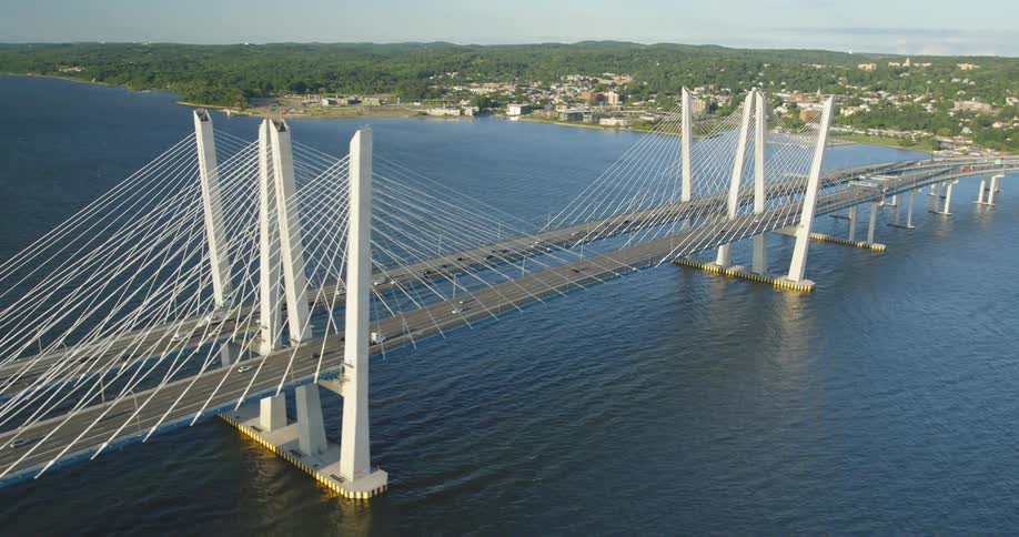 A bridge with vehicles over a river with land the sky and clouds in the background