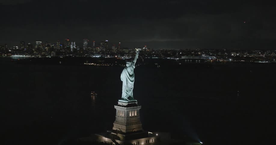 The statue of liberty is illuminated at night while the skyline of new york city is nearby