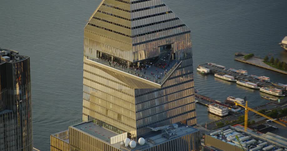 An aerial view of a steel building with a triangular balcony at the top against water with many other buildings surrounding it downtown
