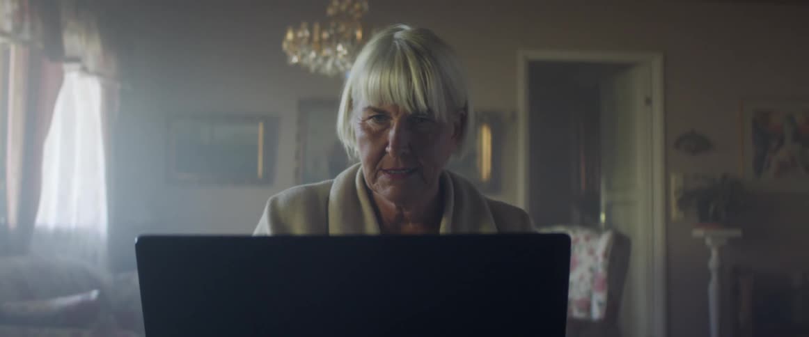 Woman with grey hair sits at a table and makes a video call on a laptop in the living room at home