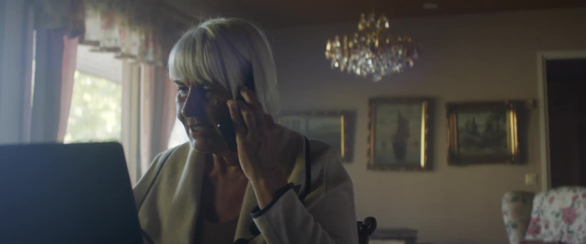 Woman with grey hair sits at a table and talks on smartphone while using laptop in living room