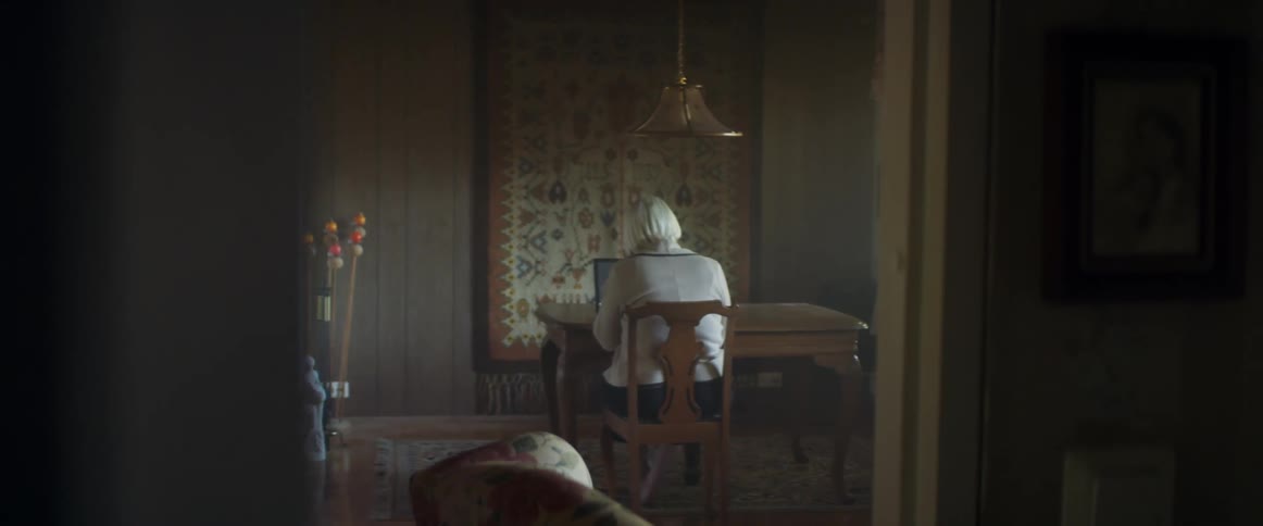Elderly woman with grey hair sits at a table uses a laptop in a room