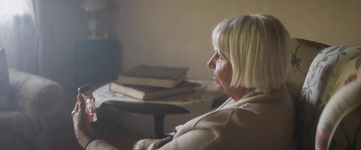 An elderly woman at home video chats on her phone with a man holding a baby