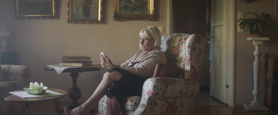 A blonde woman sits in a floral upholstered chair and smiles while talking via video call on her smartphone