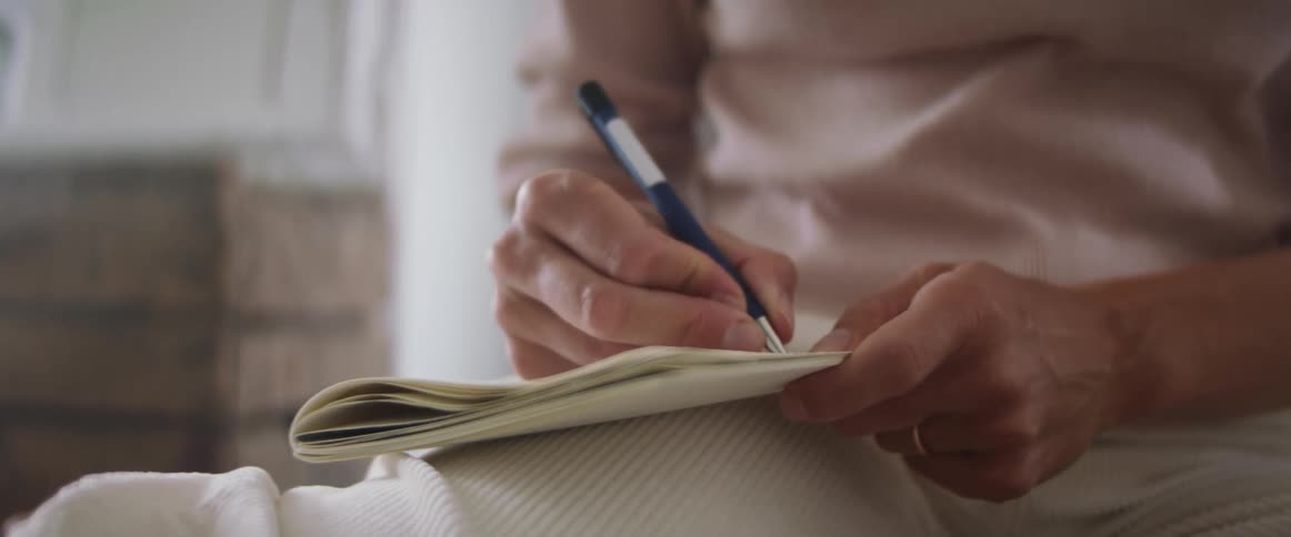 Woman with a pink sweater and white pants sitting on the ground writes things down in a notebook