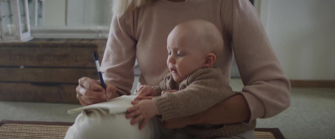 A woman sits on the floor holding her infant child and a book and pen