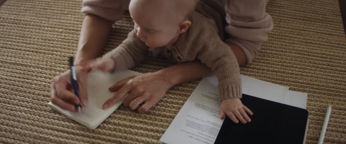 A woman rests on her elbows on the floor with a baby in her arms and tries to write on a notepad as the baby attempts to take her pen