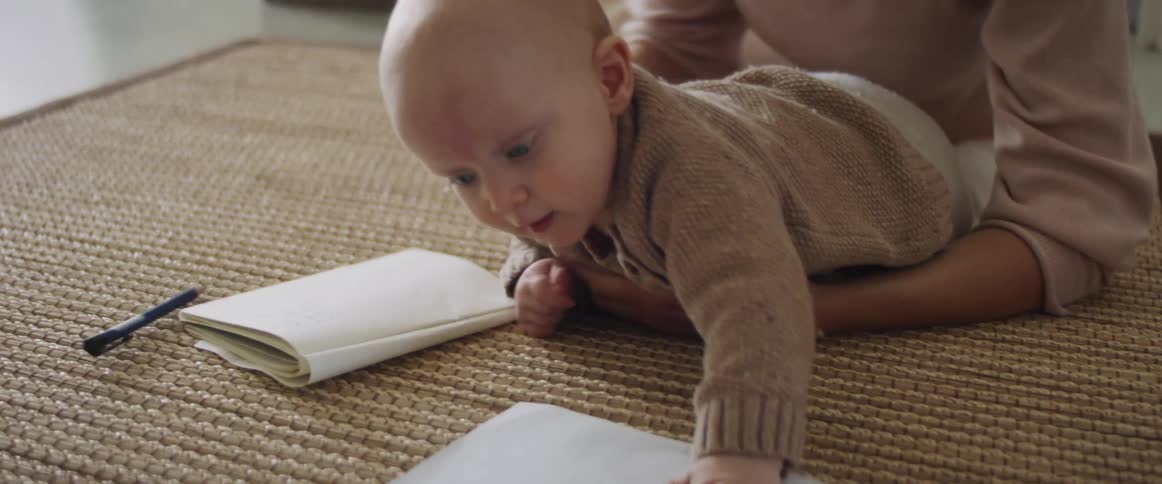 A woman lies on the floor with her baby while writing in a notebook and then sits up and picks him up
