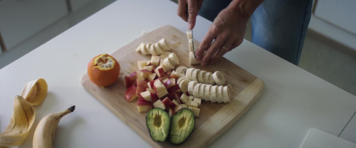 A woman with a gold wedding ring and dark skin stands at a counter chopping bananas apples avocados and oranges with blue jeans on