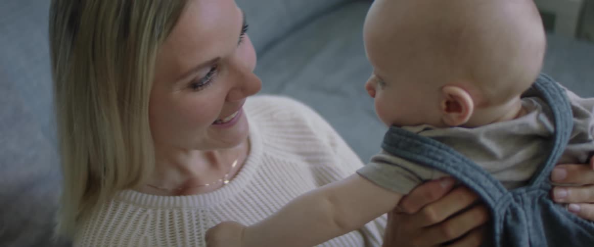 Blonde mother with a white knit shirt and sitting on a sofa holds and looks at her baby