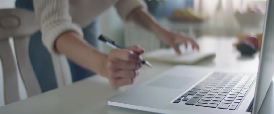 Blonde woman with a white knit sweater and jeans uses a laptop and writes things in a notebook