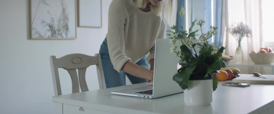 Blonde woman with a white knit sweater and jeans uses a laptop in dining room at home