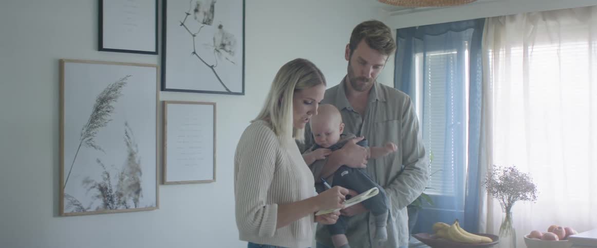 Young couple stand in a room at home with their baby talking while taking notes on a notebook