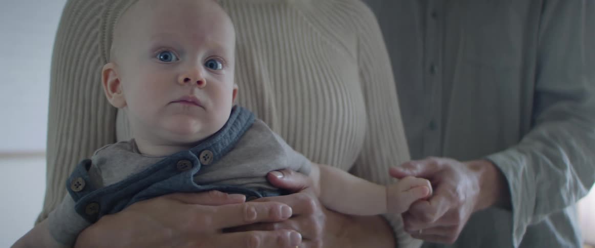 A baby turns to look into the camera as he is held by a woman who is in turn being embraced by a man