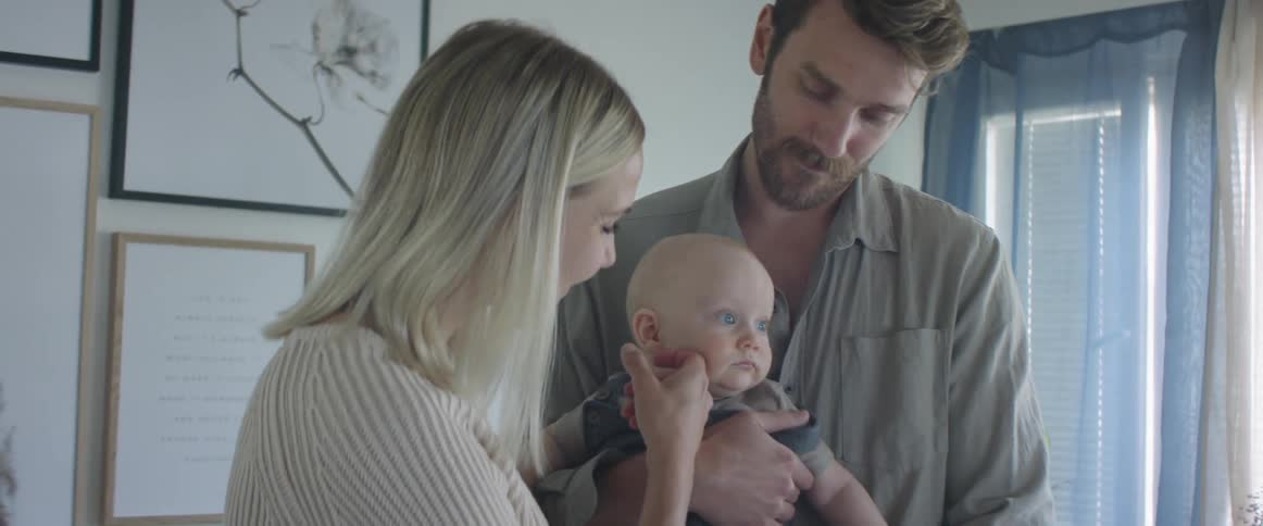 A man in his house holds his baby son and kisses his head as his wife rubs the babys cheek