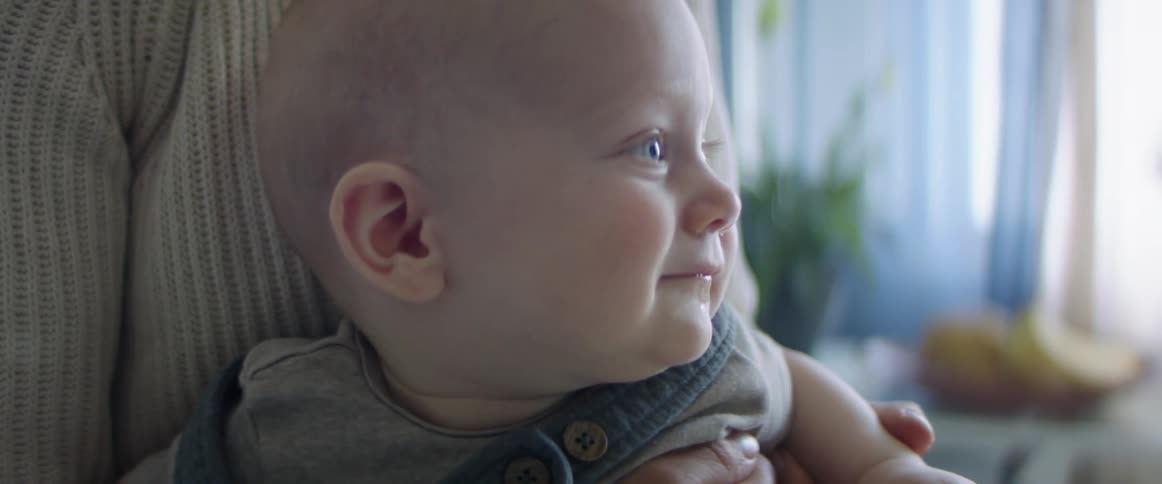 A woman holding a baby with a wall a plant and a window in the background