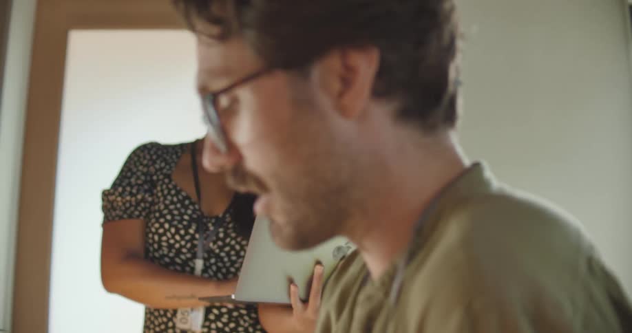 A woman holding a silver apple laptop stands next to a man sitting down and talking inside of an office room at daytime