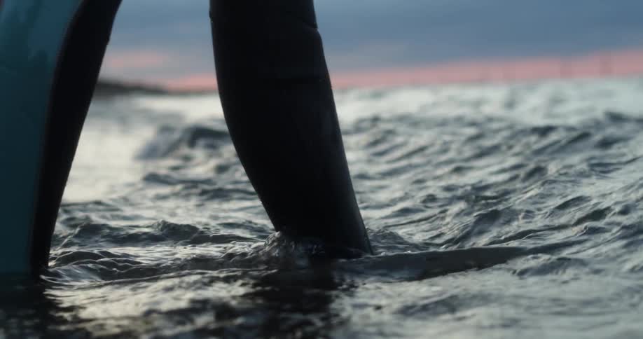 A person wearing a wetsuit steps into the shallow sea waters on the coast at sunset